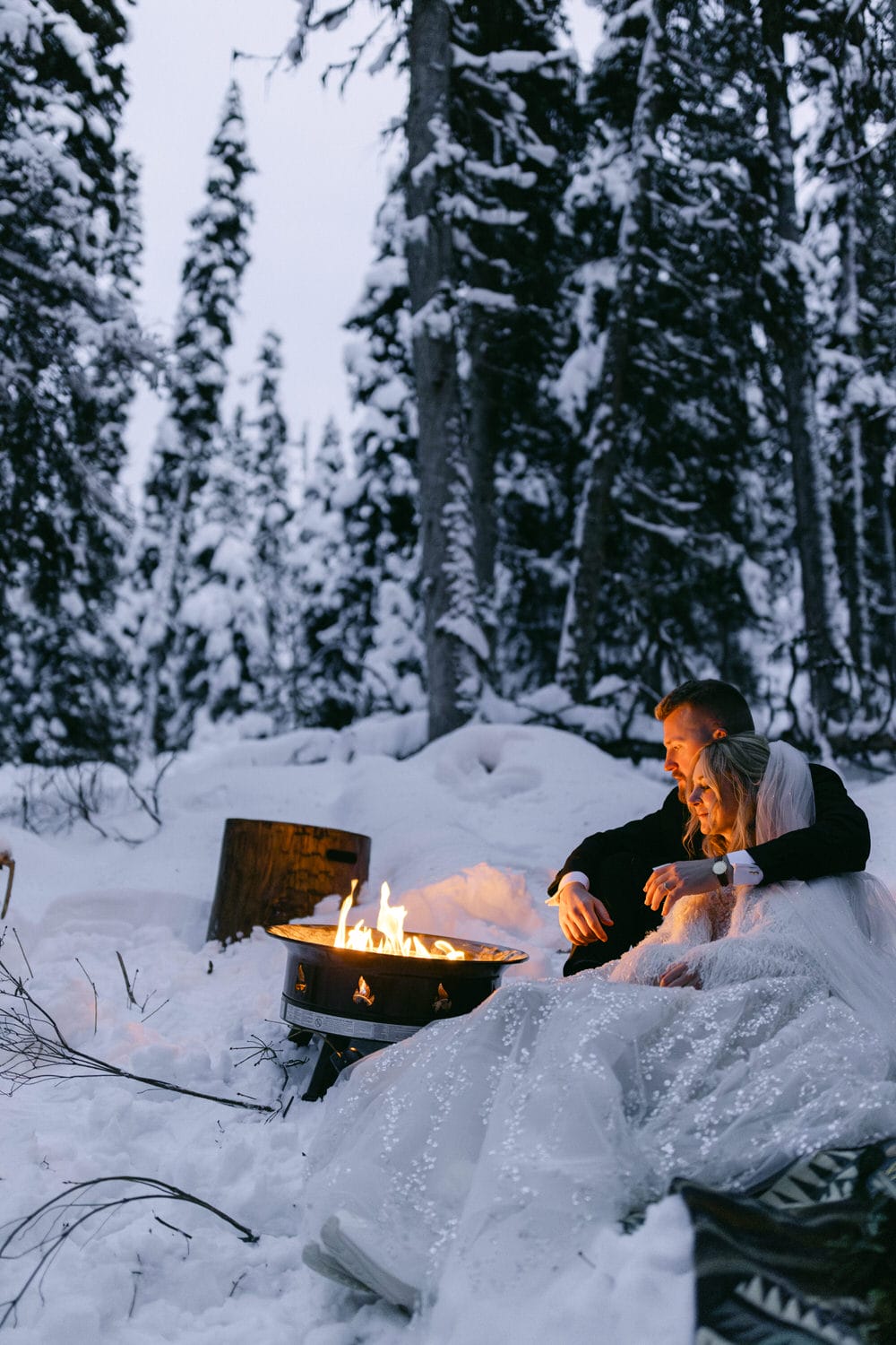 A couple in wedding attire cuddling next to a fire pit in a snowy forest setting