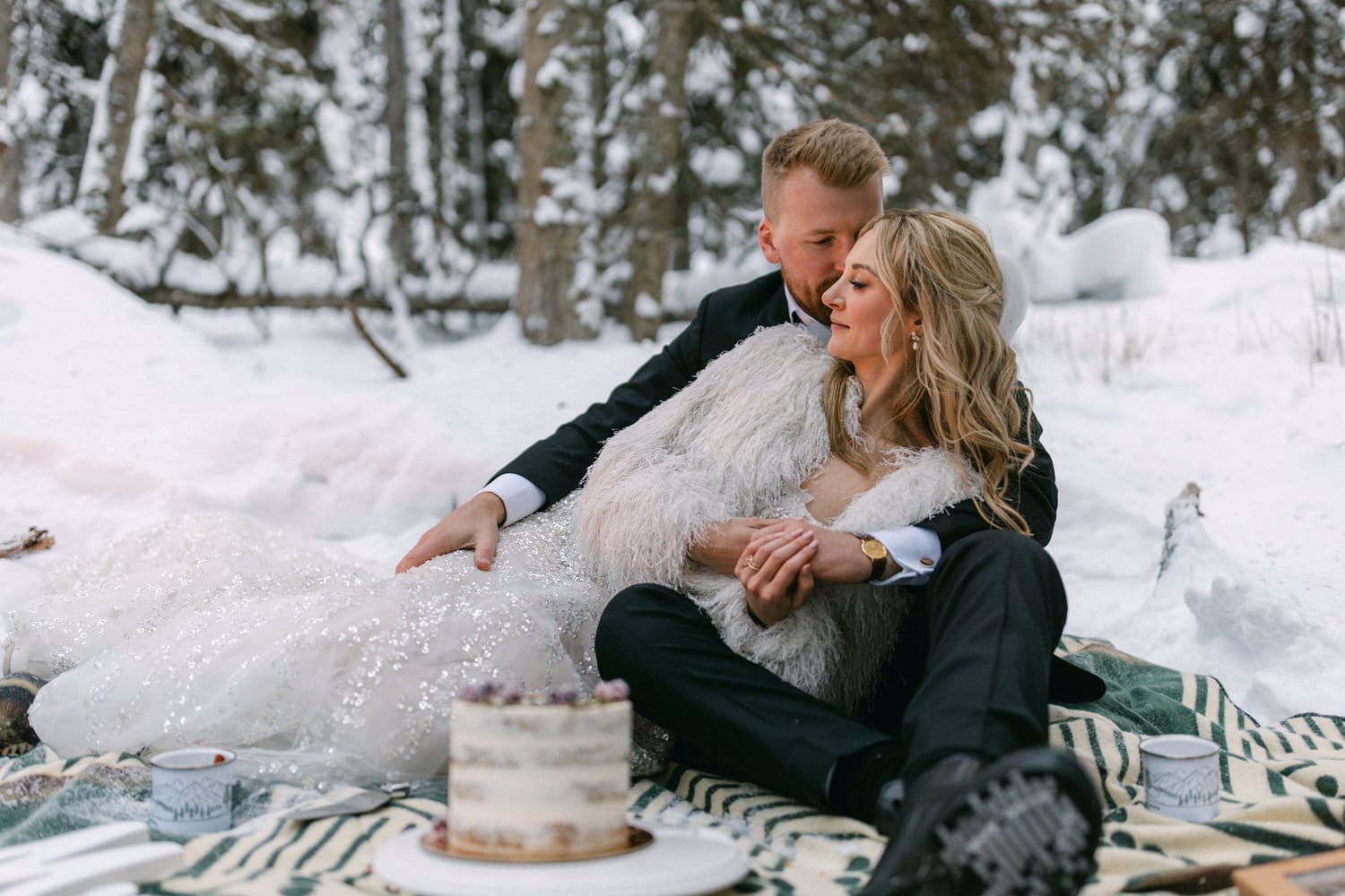 A bride and groom in elegant attire sitting closely on a blanket in a snowy landscape, with a small cake and winter trees in the background.