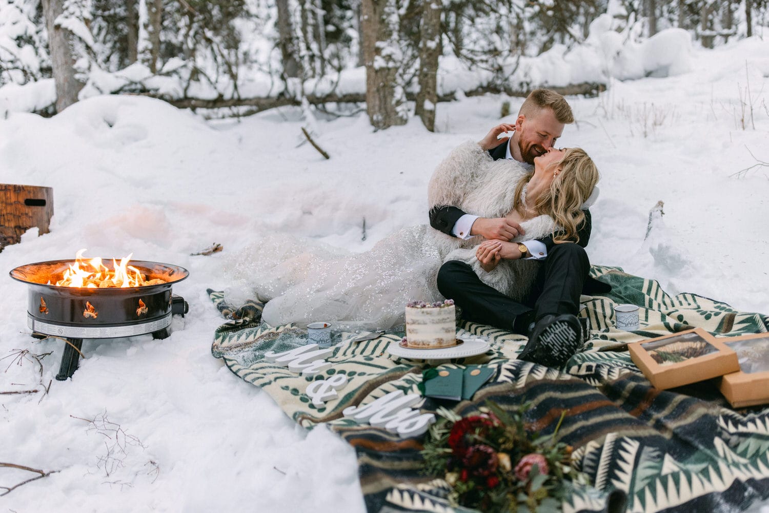 A couple in wedding attire cozying up beside a fire pit in a snowy setting with a "Mr. & Mrs." sign and wedding cake nearby.