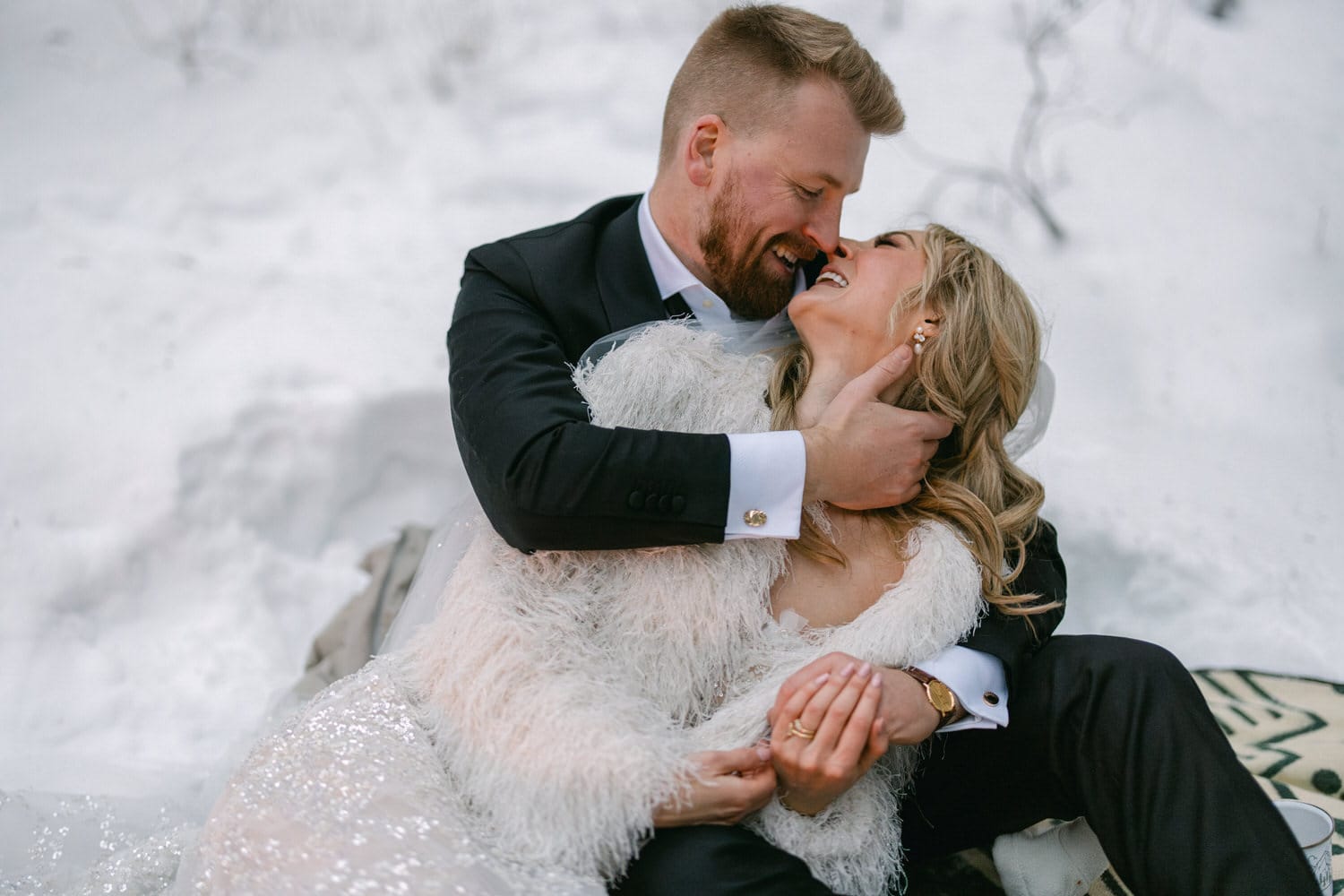 A joyful couple embracing and laughing in a snowy setting, with the bride in a white dress with a furry shawl and the groom in a black suit.