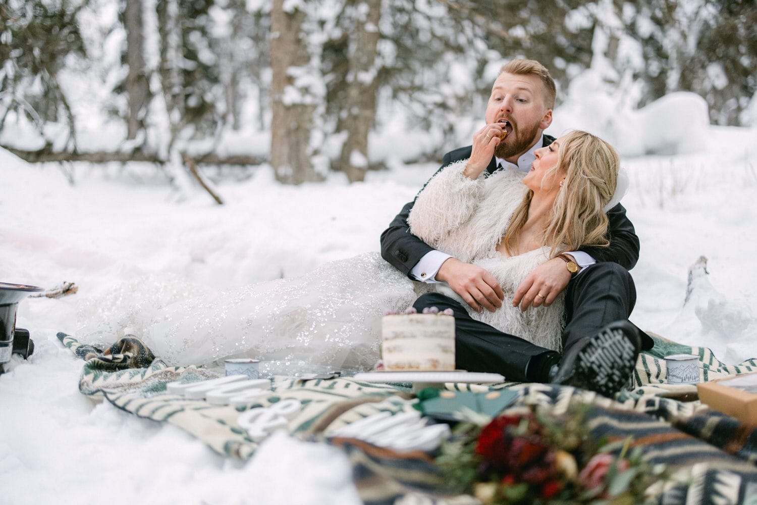 A bride and groom sitting on a blanket in the snow, sharing cake and enjoying a moment together.