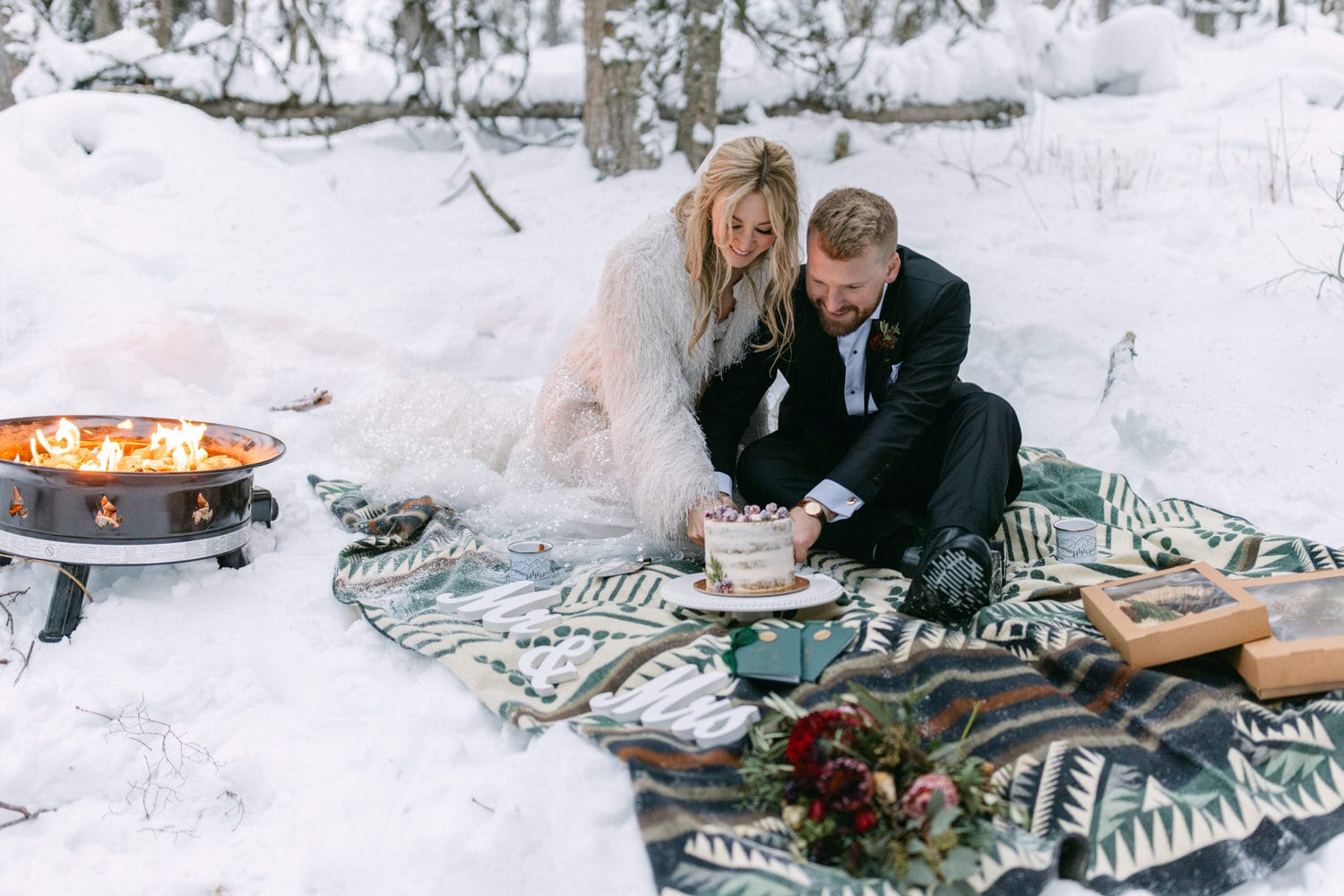 A couple in wedding attire cutting a cake outdoors on a snow-covered ground with a fire pit nearby and winter-themed decorations.