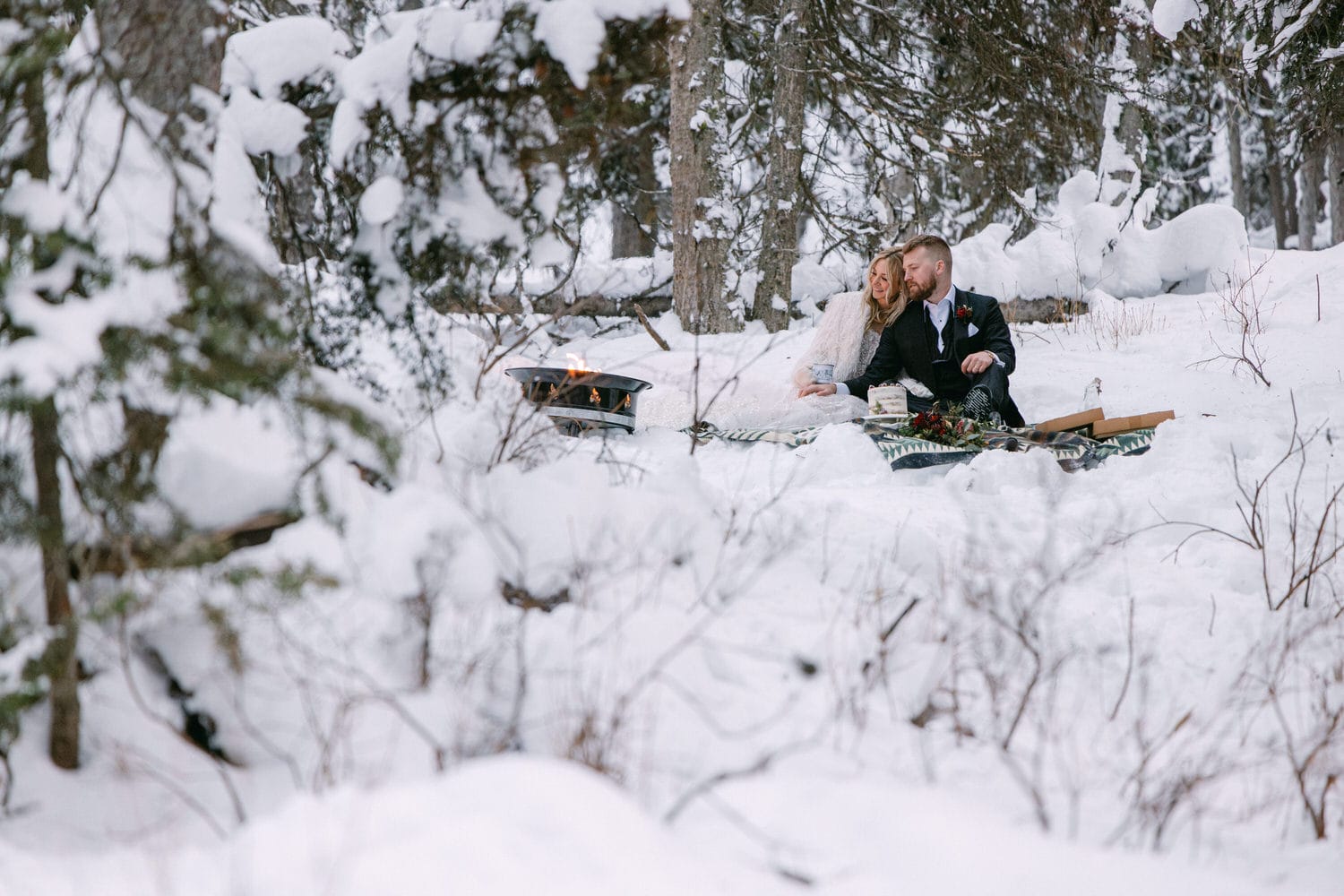 A couple enjoys a romantic picnic surrounded by a snowy forest landscape, with a small fire pit adding warmth to the serene winter setting.