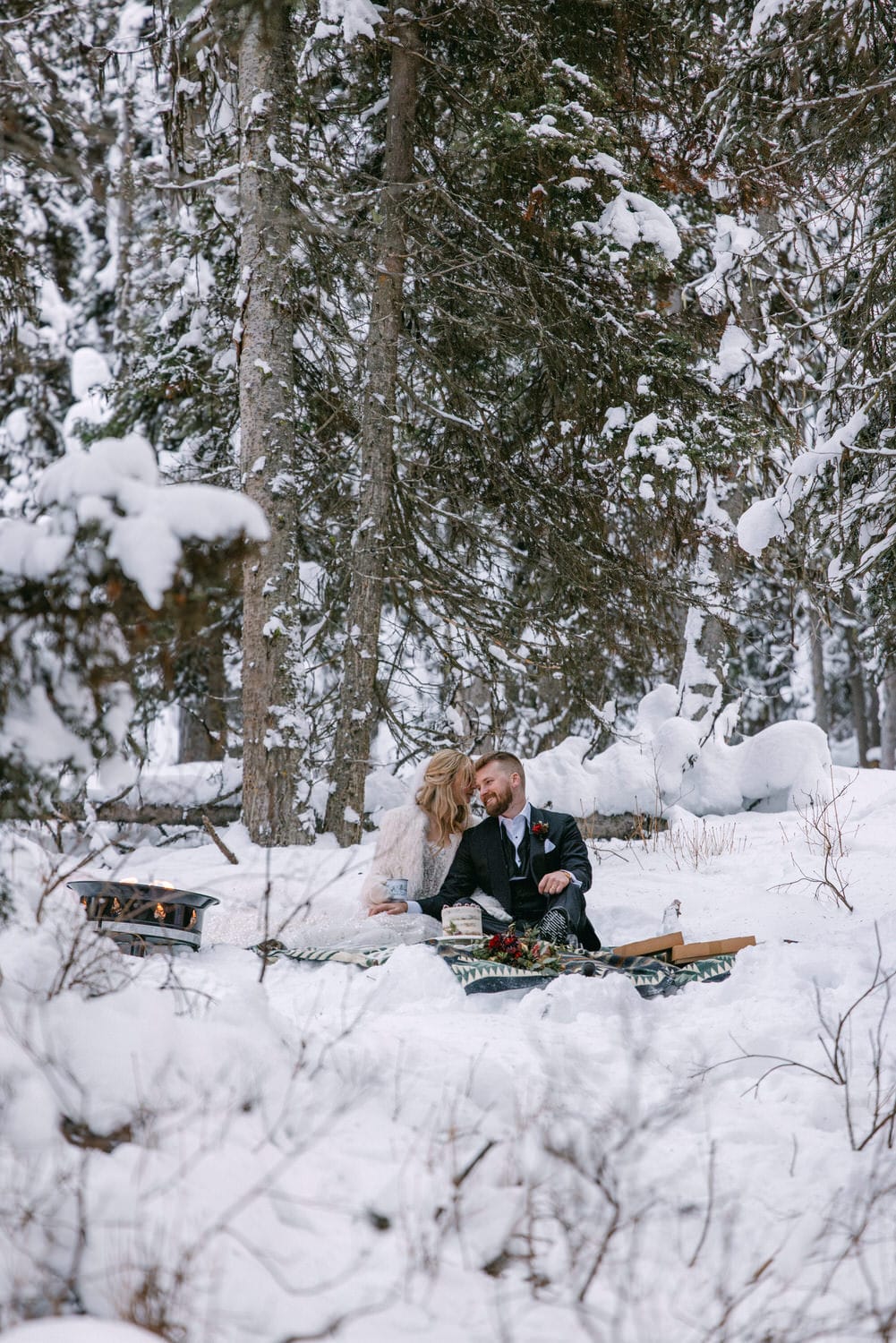 A couple in wedding attire having a romantic moment on a snowy forest backdrop with a fire pit nearby.