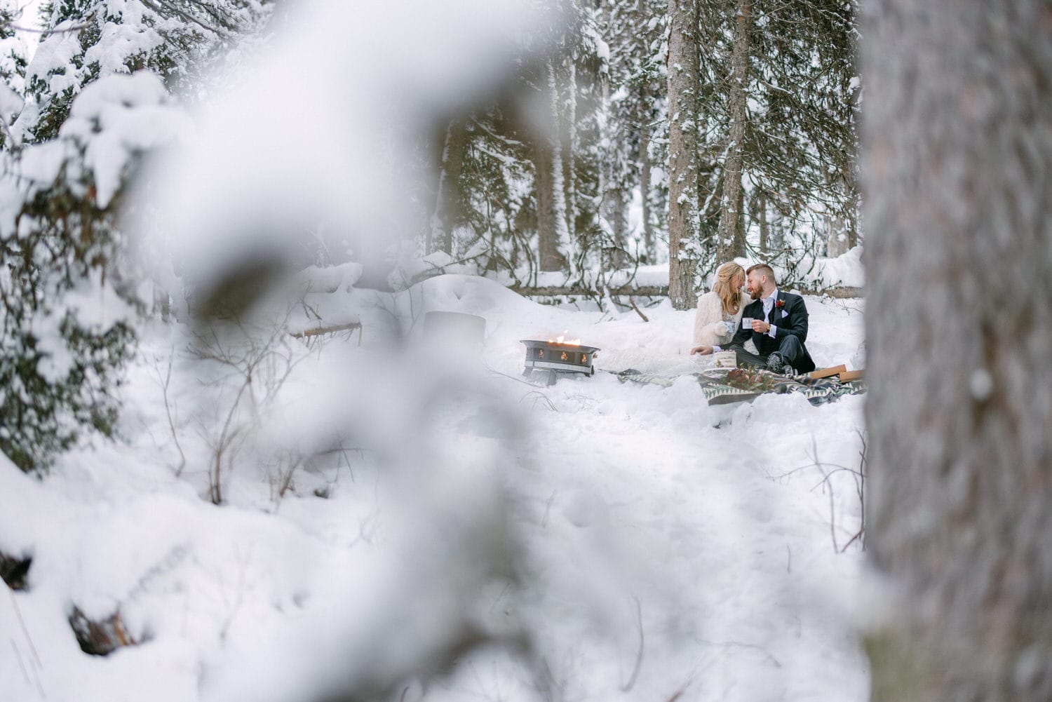 Two people enjoying a romantic picnic in a snowy forest with a portable fireplace.