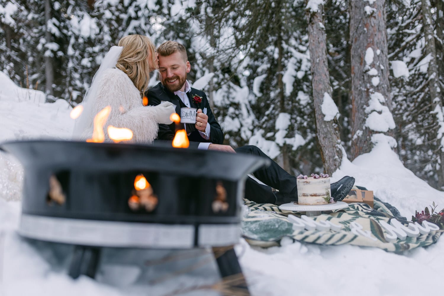 Winter Wedding Romance by the Fire:::A couple in wedding attire sharing a romantic moment by a fire pit in a snowy setting, with a cake and "love" sign visible.