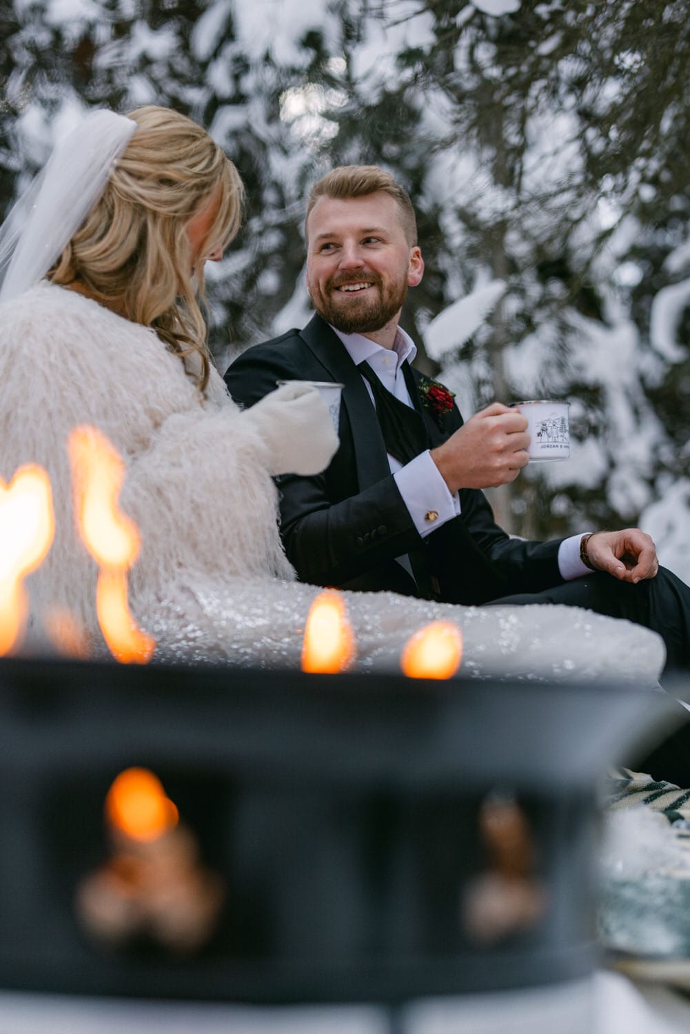 A bride and groom holding cups and making a toast, with a focus on the smiling groom, as candles and snow add romance to the scene.