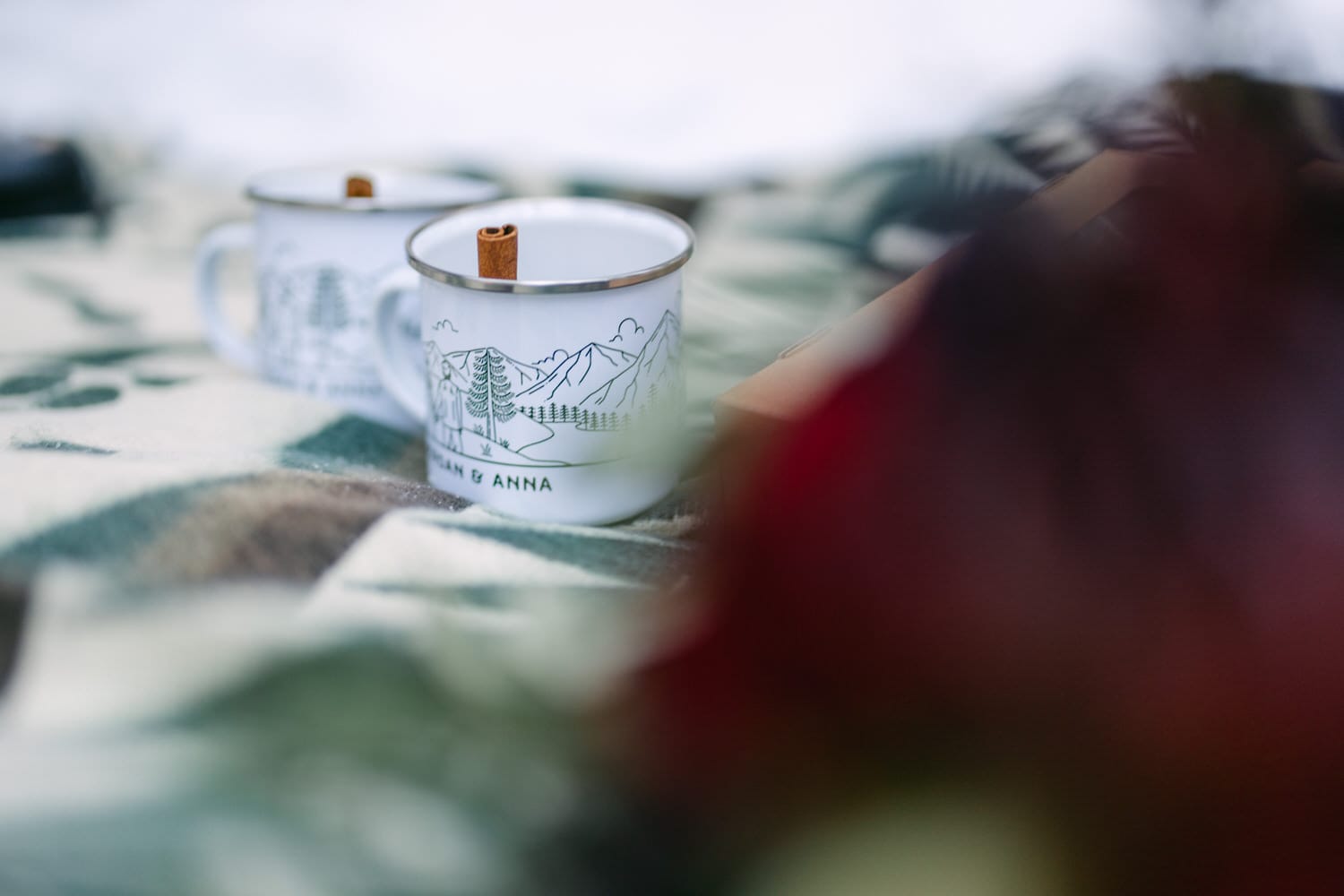 Two enamel camping mugs with mountain design and cinnamon sticks on a plaid blanket with soft focus foreground