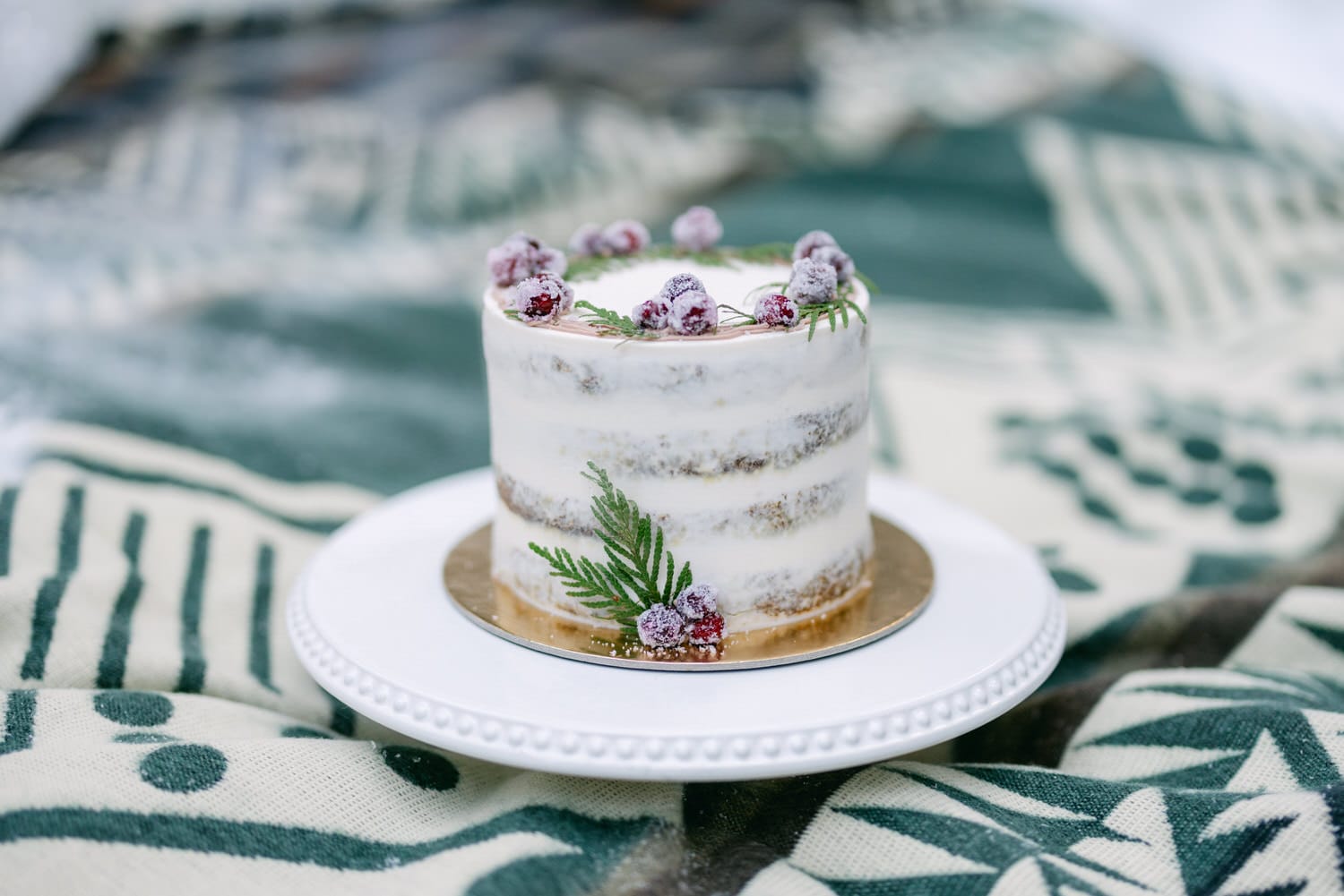 A small naked cake with white frosting, decorated with sugared berries and greenery, presented on a white plate atop a green and cream patterned cloth.