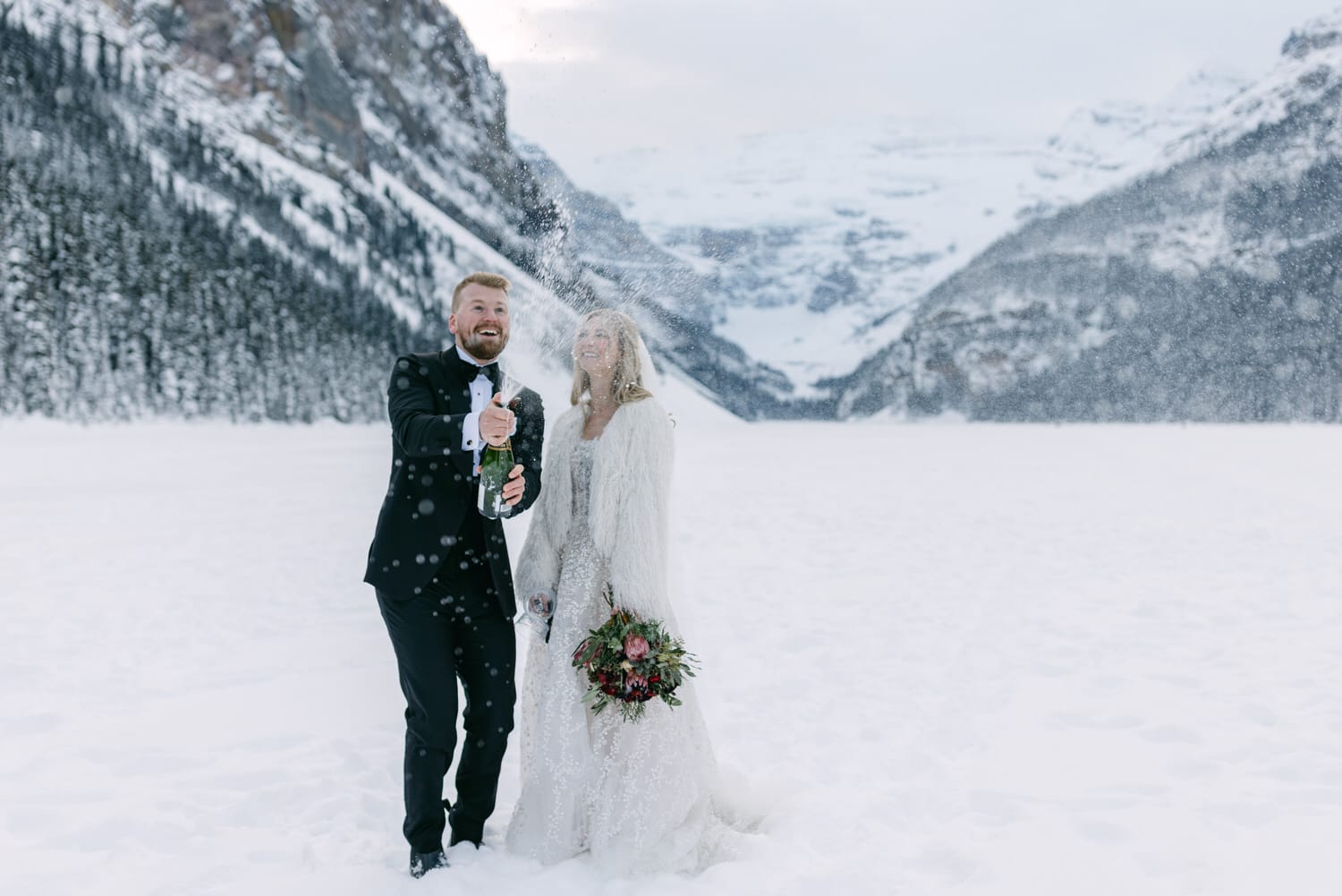 A bride and groom celebrating with champagne in a snowy landscape with mountains in the background.