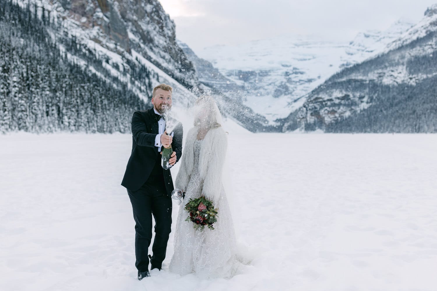 A bride and groom celebrating by opening a bottle of champagne in a snowy mountain landscape