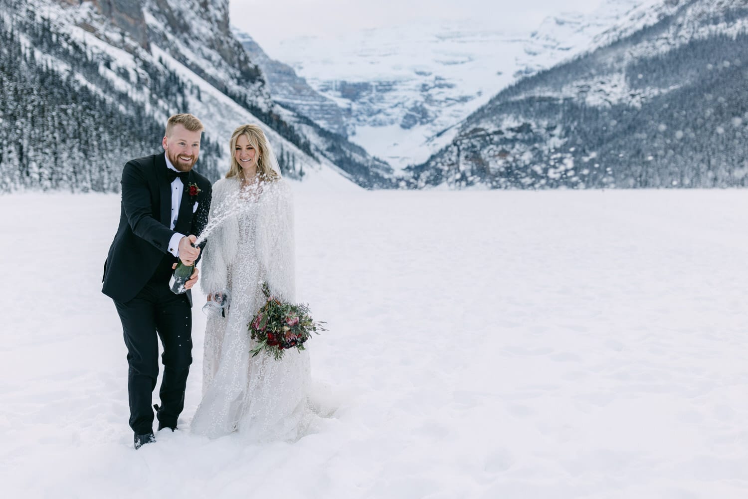 A couple in wedding attire celebrating with a champagne spray in a snowy mountain landscape.