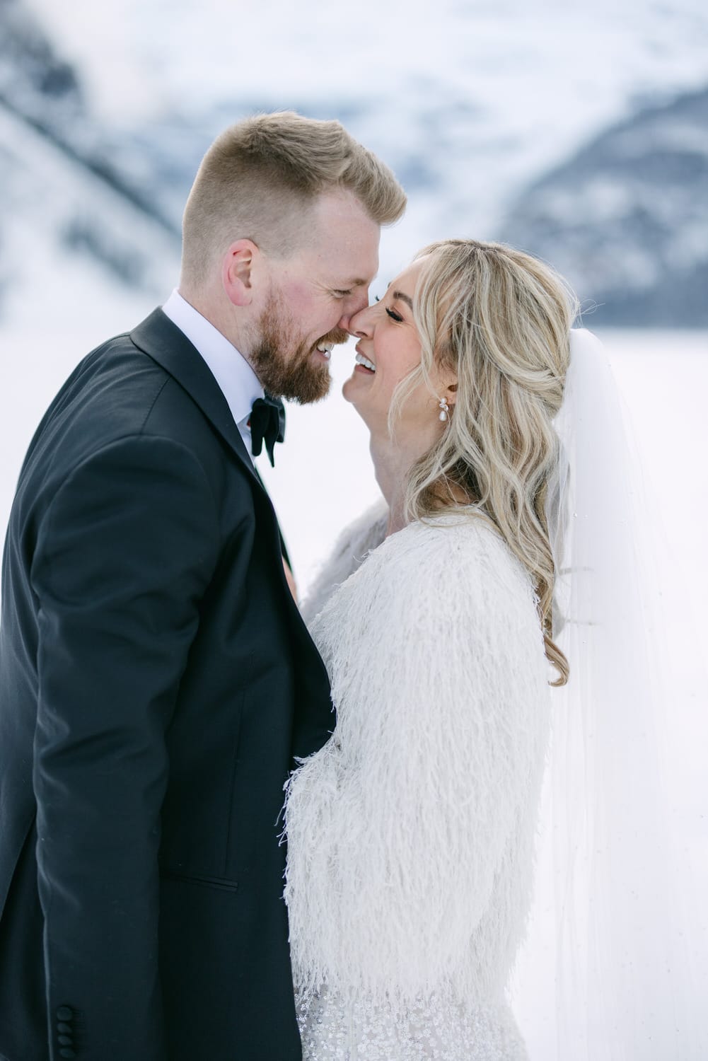 A bride and groom smiling at each other in a snowy landscape, the bride in a white dress with a fuzzy jacket and the groom in a black suit.