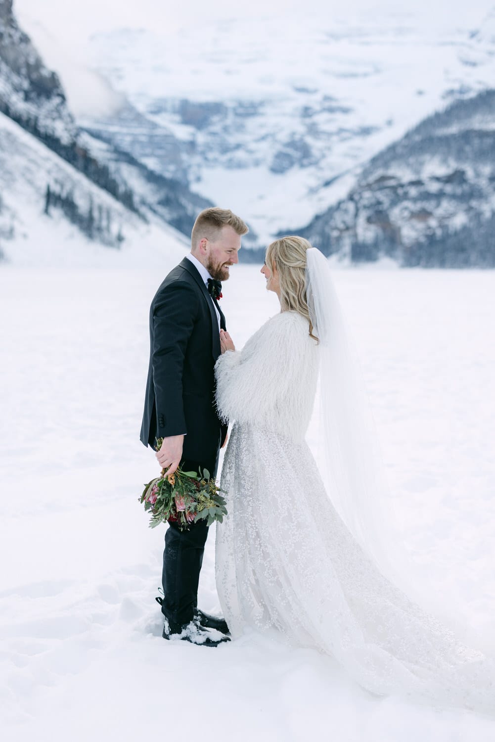 A bride and groom sharing an intimate moment in a snowy landscape, with the bride holding a bouquet and both dressed in formal wedding attire.