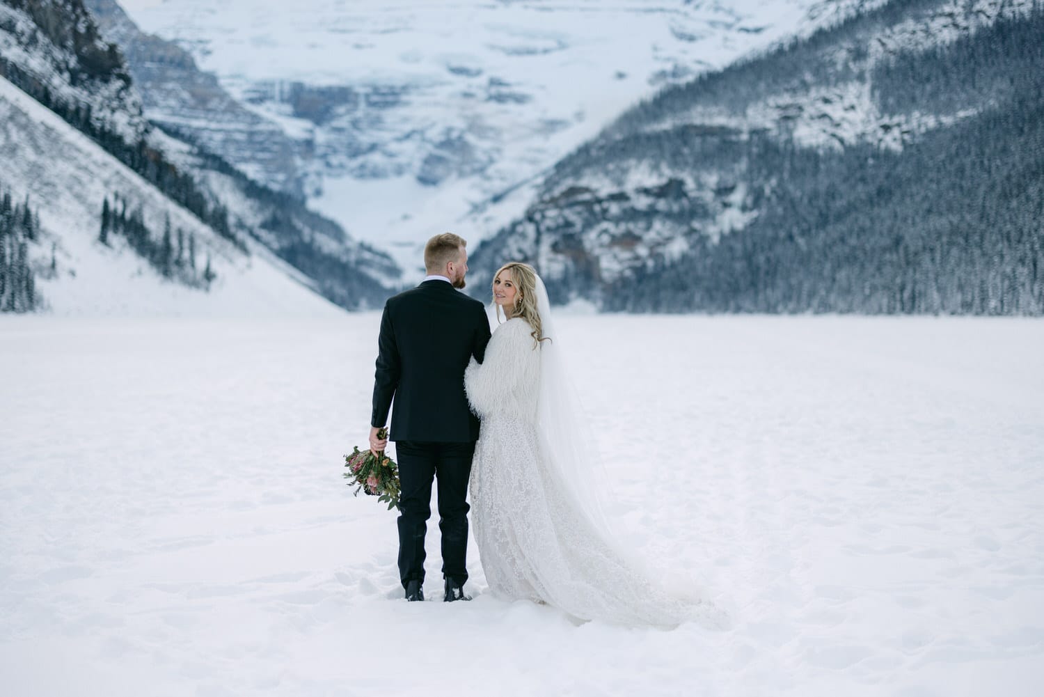 A bride and groom holding hands in a snow-covered landscape with mountain backdrop.