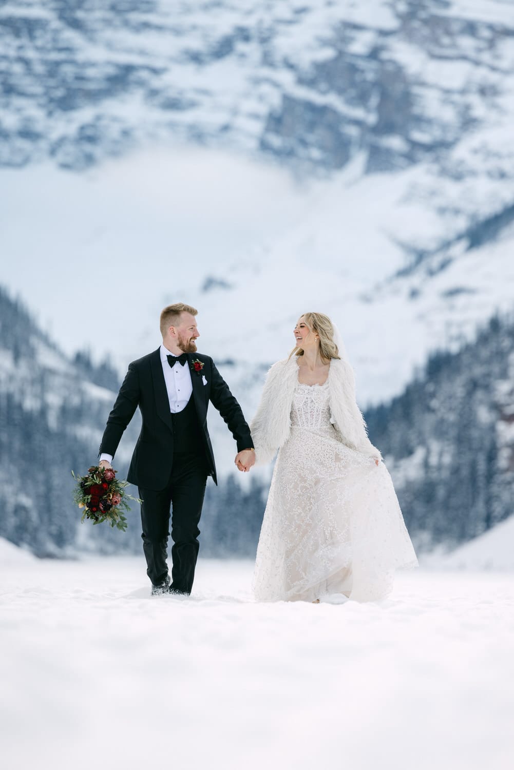 Bride and groom holding hands and smiling at each other in a snowy landscape with mountains in the background.