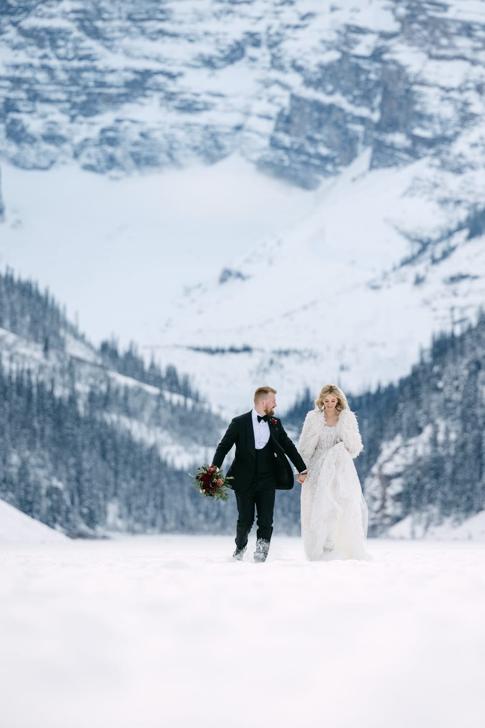 A bride and groom walking hand in hand in a snowy landscape with mountains in the background.