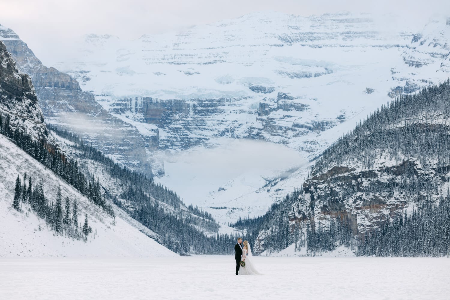 A couple in wedding attire holding hands in a vast snowy landscape with towering, snow-covered mountains in the background.