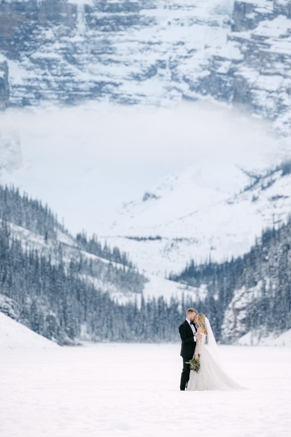 A bride and groom embracing in a snowy landscape with snow-covered mountains in the background.