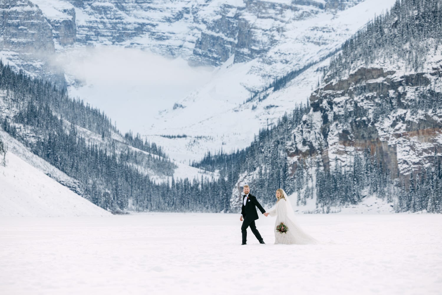 A bride and groom holding hands in a vast snowy landscape with forested mountains in the background.