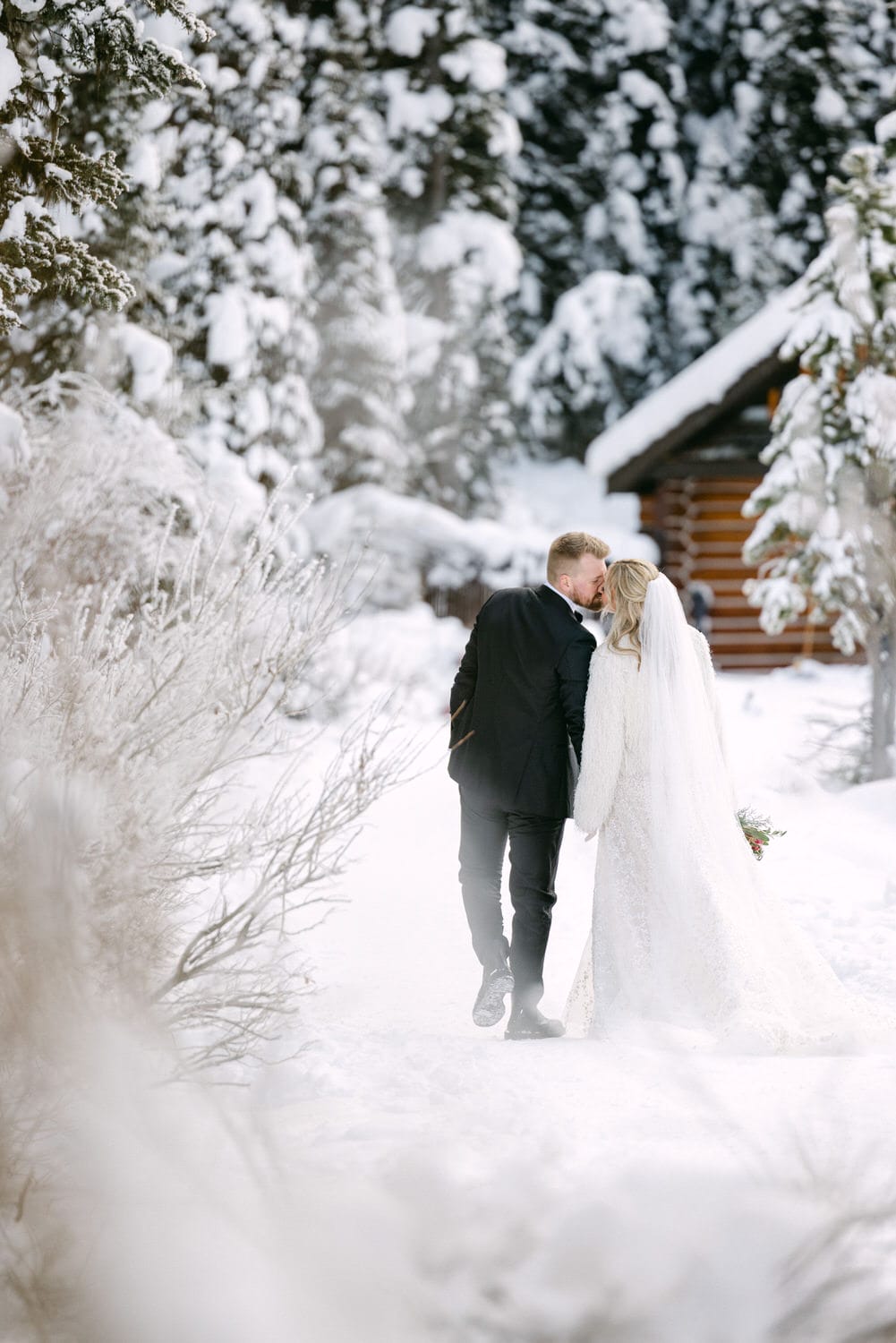 A bride and groom sharing a kiss on a snowy path with evergreen trees and a wooden cabin in the background.