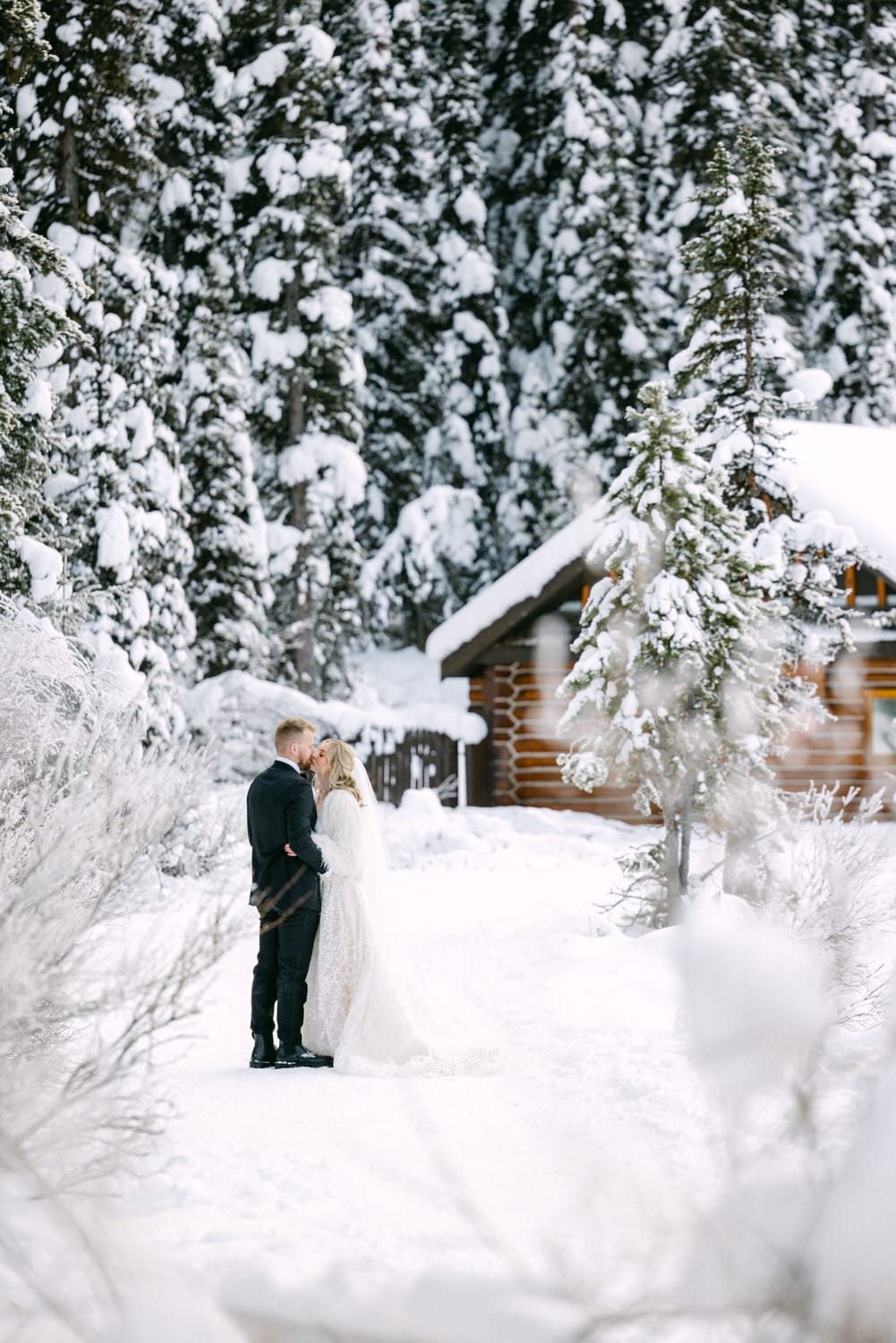 A couple embracing surrounded by a snowy winter landscape with snow-covered trees and a wooden cabin in the background.