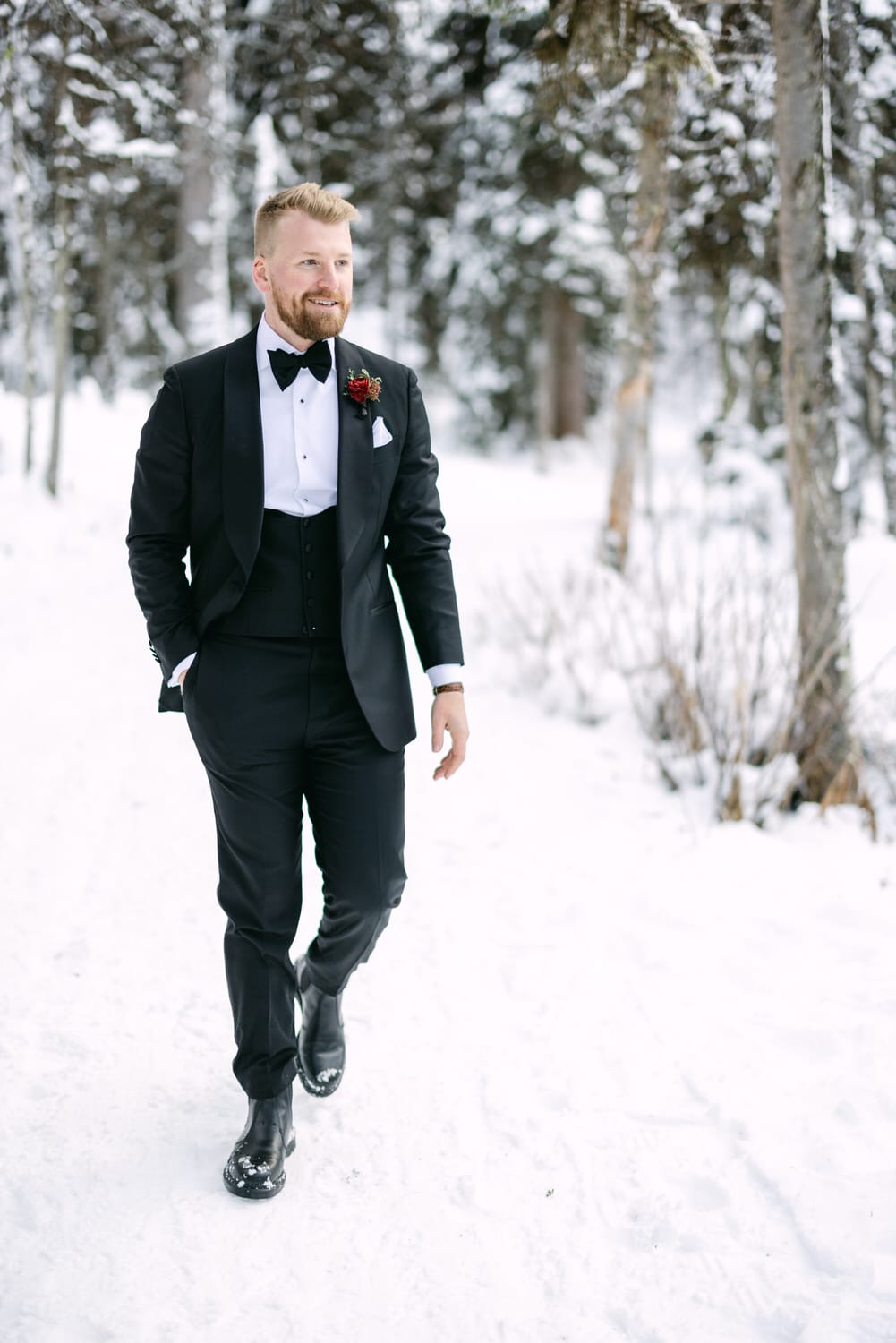 A man in a black tuxedo with a boutonniere walking confidently in the snow, with a background of snowy trees.