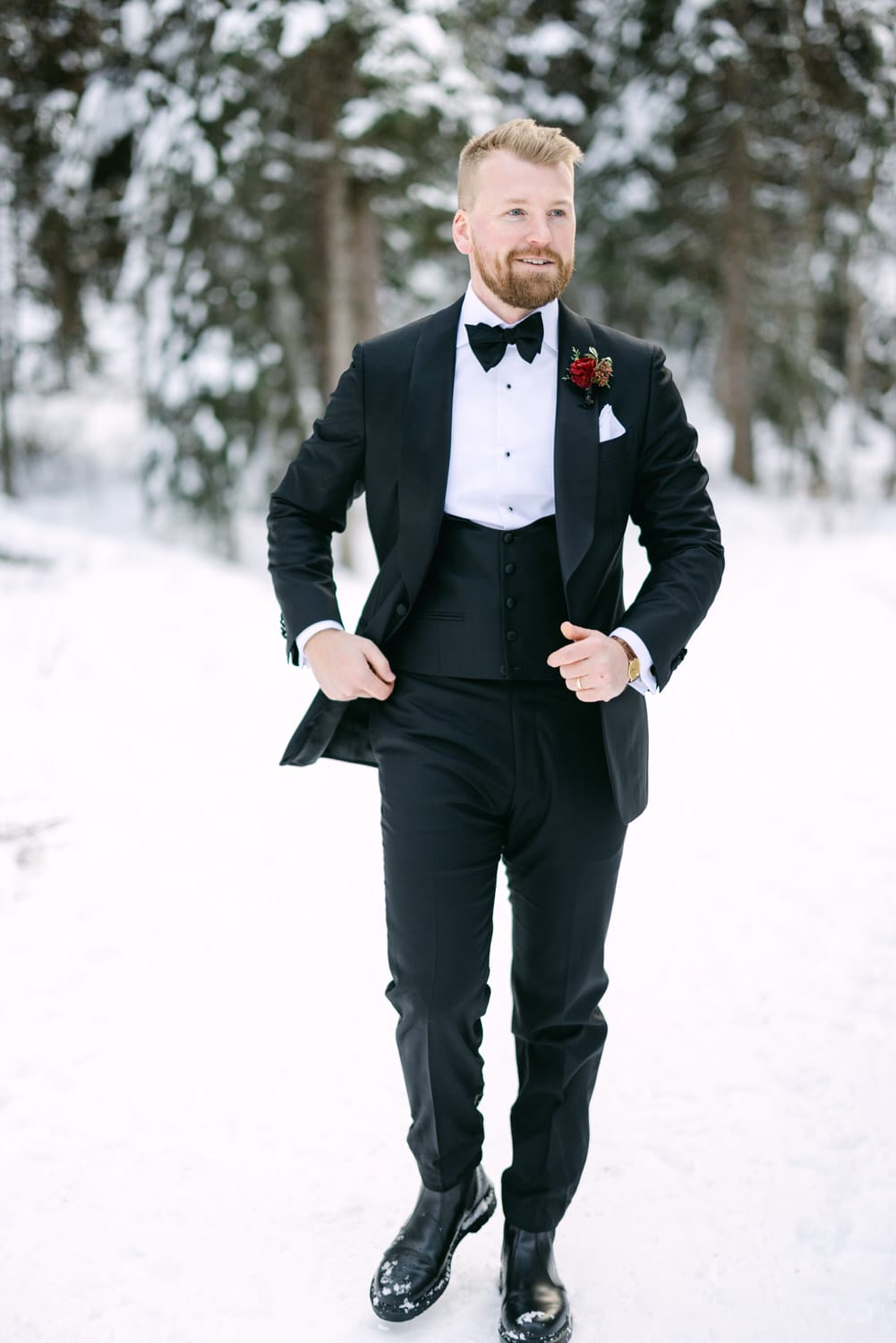 A man dressed in a black suit with a bow tie and pocket square, wearing a boutonniere, standing in a snowy landscape with trees in the background