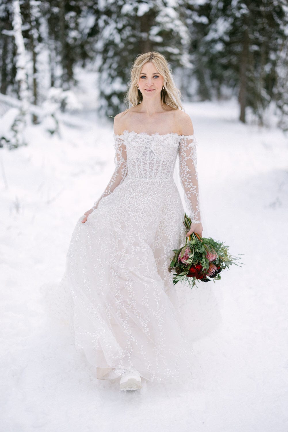 A woman in a white, off-the-shoulder wedding dress holding a bouquet, standing in a snowy forest setting.