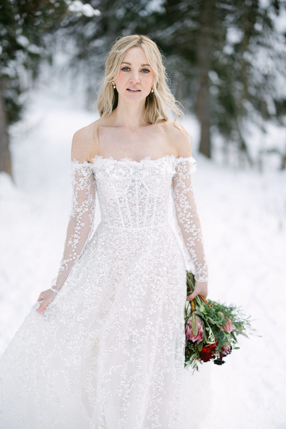 A woman in an elegant off-the-shoulder wedding dress holding a bouquet, with snowy forest background.