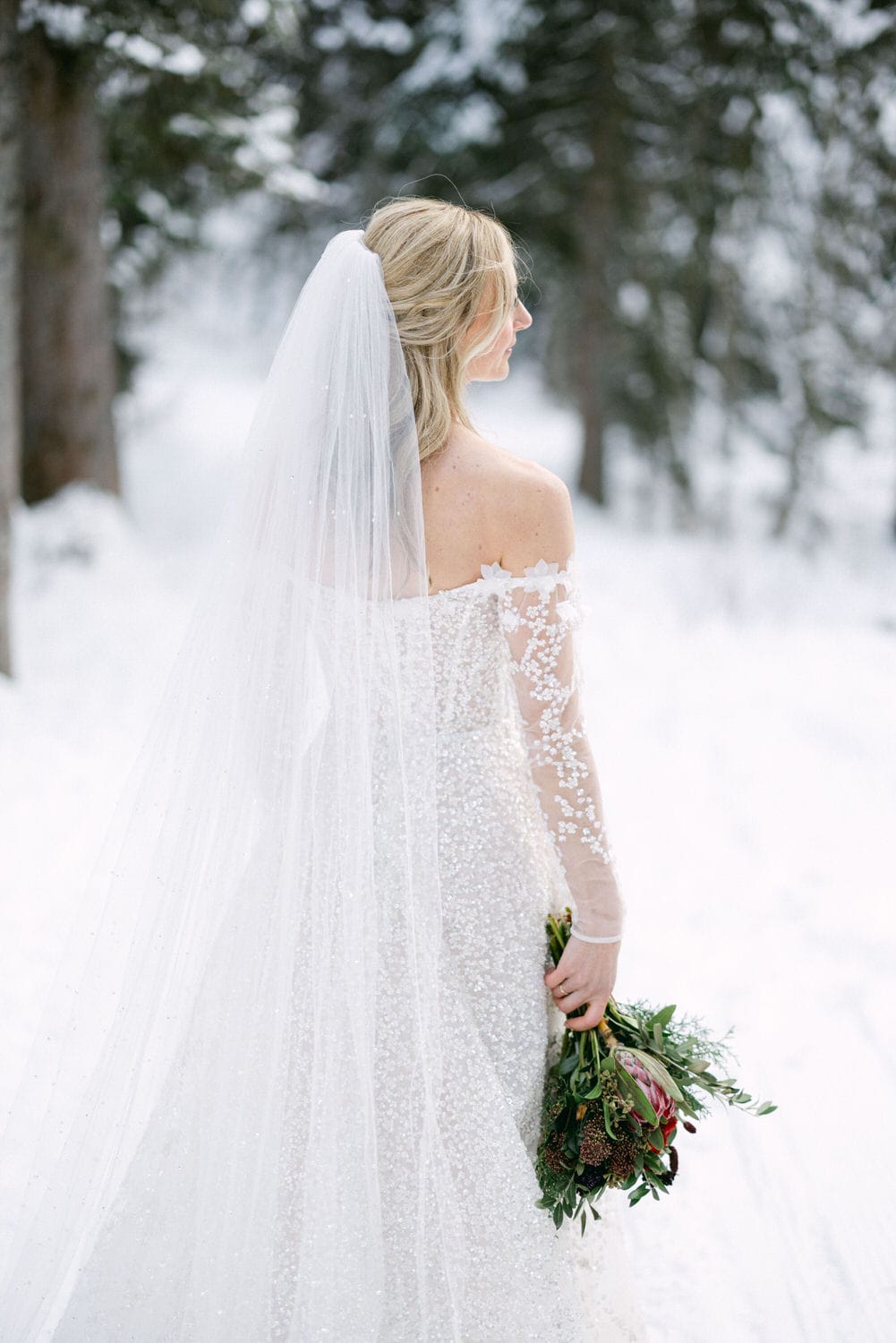 A bride in a beautiful white gown and veil holding a bouquet, poised elegantly against a snowy forest backdrop.