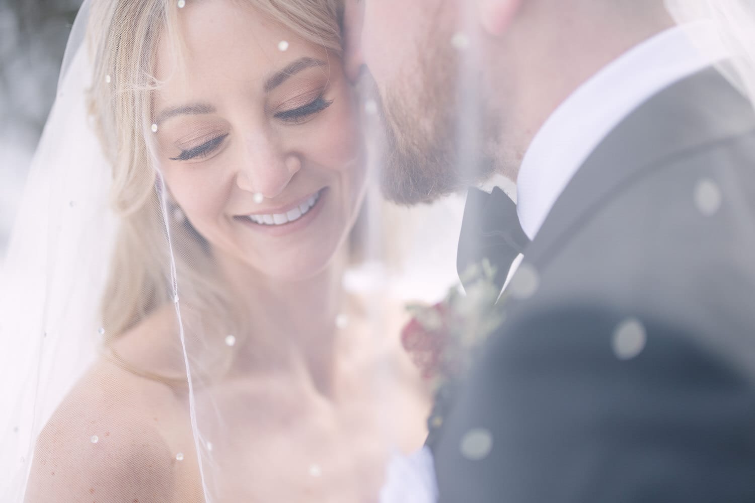 Close-up of a bride smiling under her veil with snowflakes around, groom partially visible in foreground.