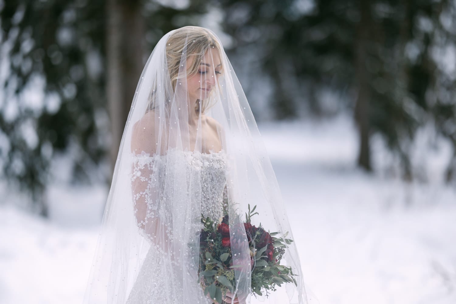 A bride in a detailed wedding gown and veil holding a bouquet, standing contemplatively amidst a snowy landscape.