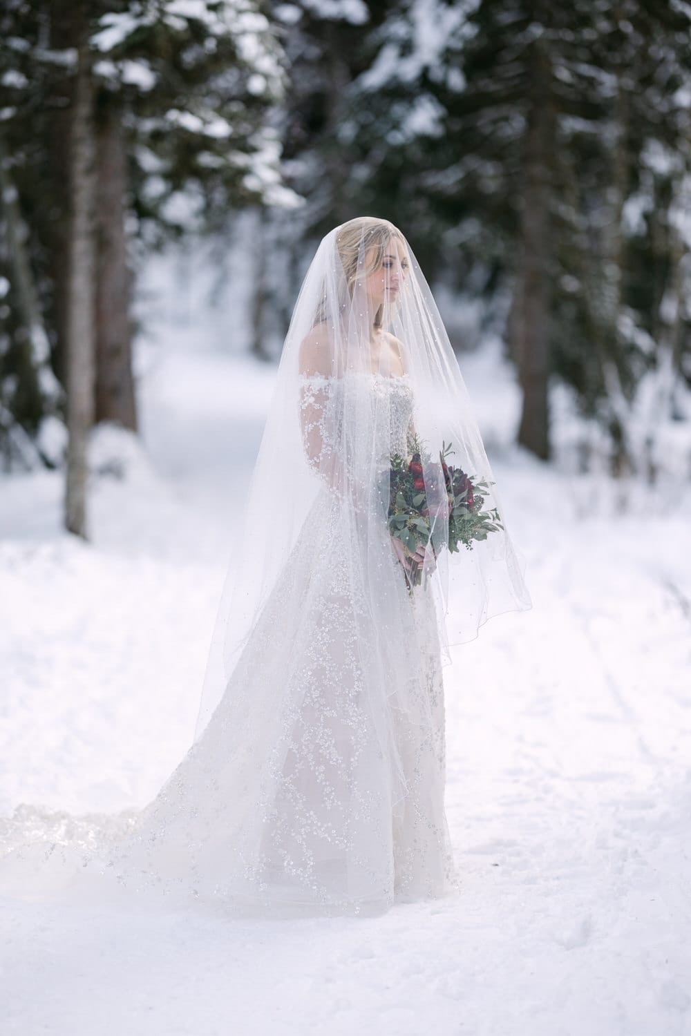 A bride in a white dress holding a bouquet standing in a snowy forest.