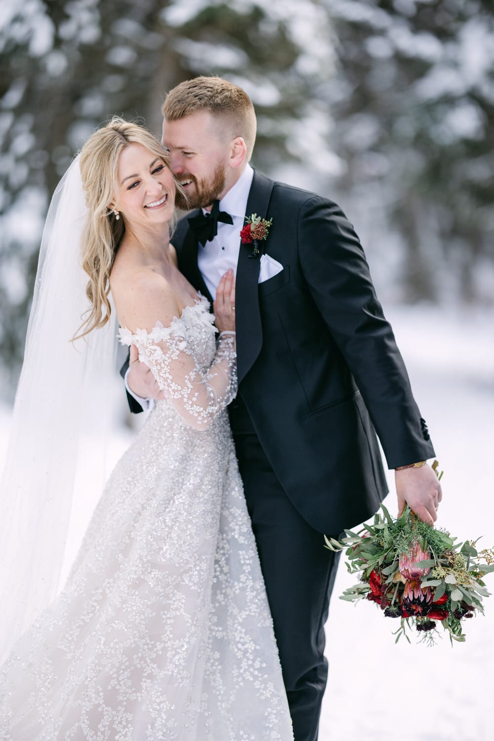 A bride and groom embracing in a snowy landscape, the bride in a lace wedding gown and the groom in a black suit, both smiling happily.