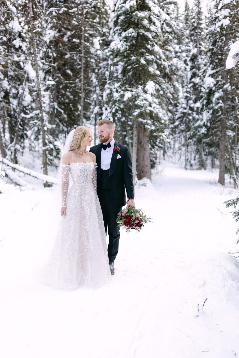 A bride and groom holding hands while walking in a snowy forest, both dressed in wedding attire.