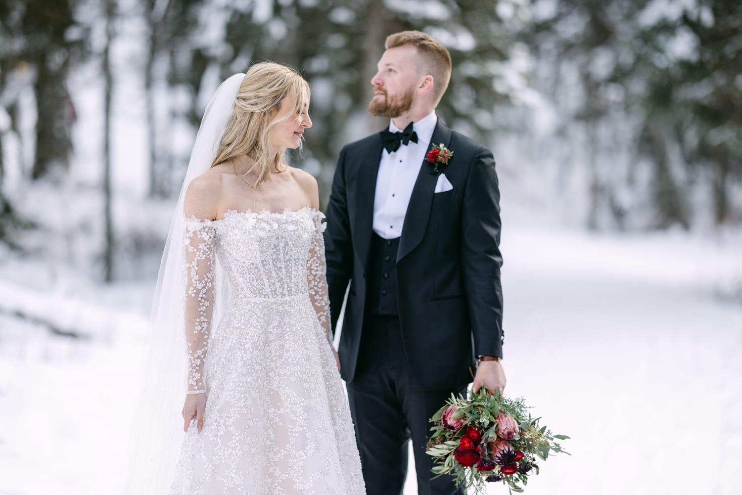 Bride in an embroidered white dress and groom in a black tuxedo looking at each other affectionately in a snowy forest setting