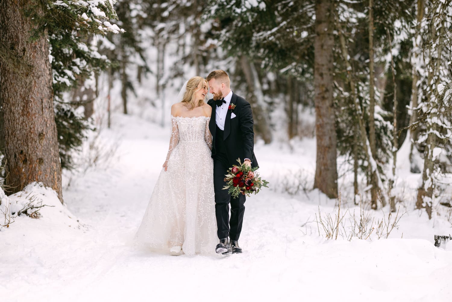 A bride in a white gown and a groom in a black suit share an affectionate moment in a snowy forest setting.