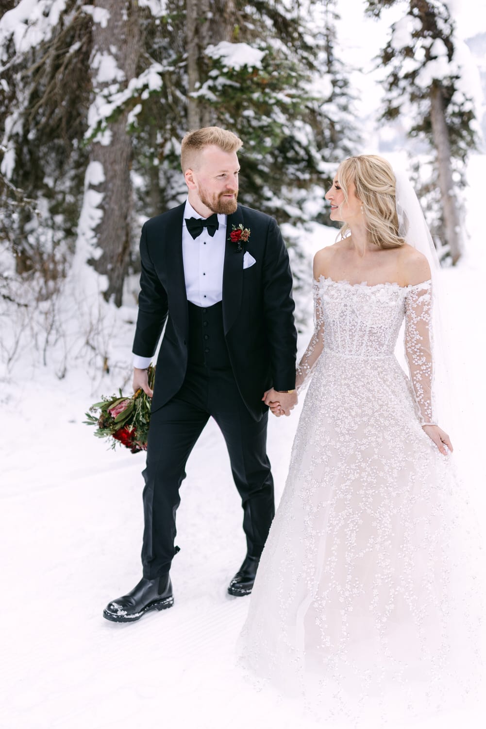 A bride and groom holding hands in a snowy forest scene, both dressed in formal wedding attire with the bride in a white lace gown and the groom in a black tuxedo with a bow tie.