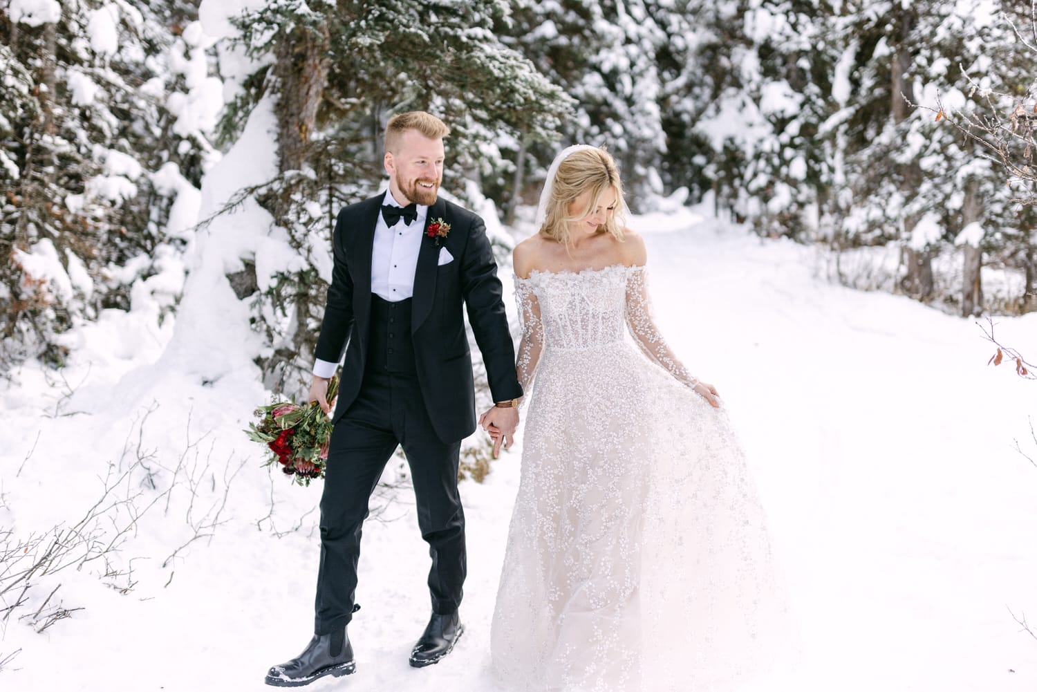 A bride in a detailed white gown and groom in a black tuxedo walking hand in hand in a snowy forest setting.