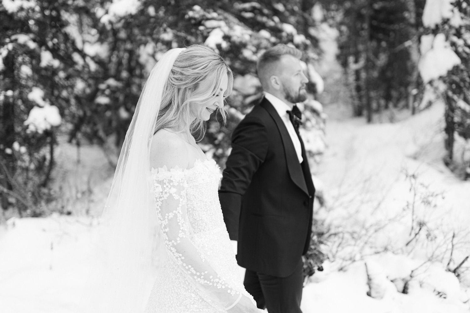 A bride and groom standing in a snowy landscape, the bride in focus with a joyful expression, and the groom slightly out of focus looking away, both in elegant wedding attire.