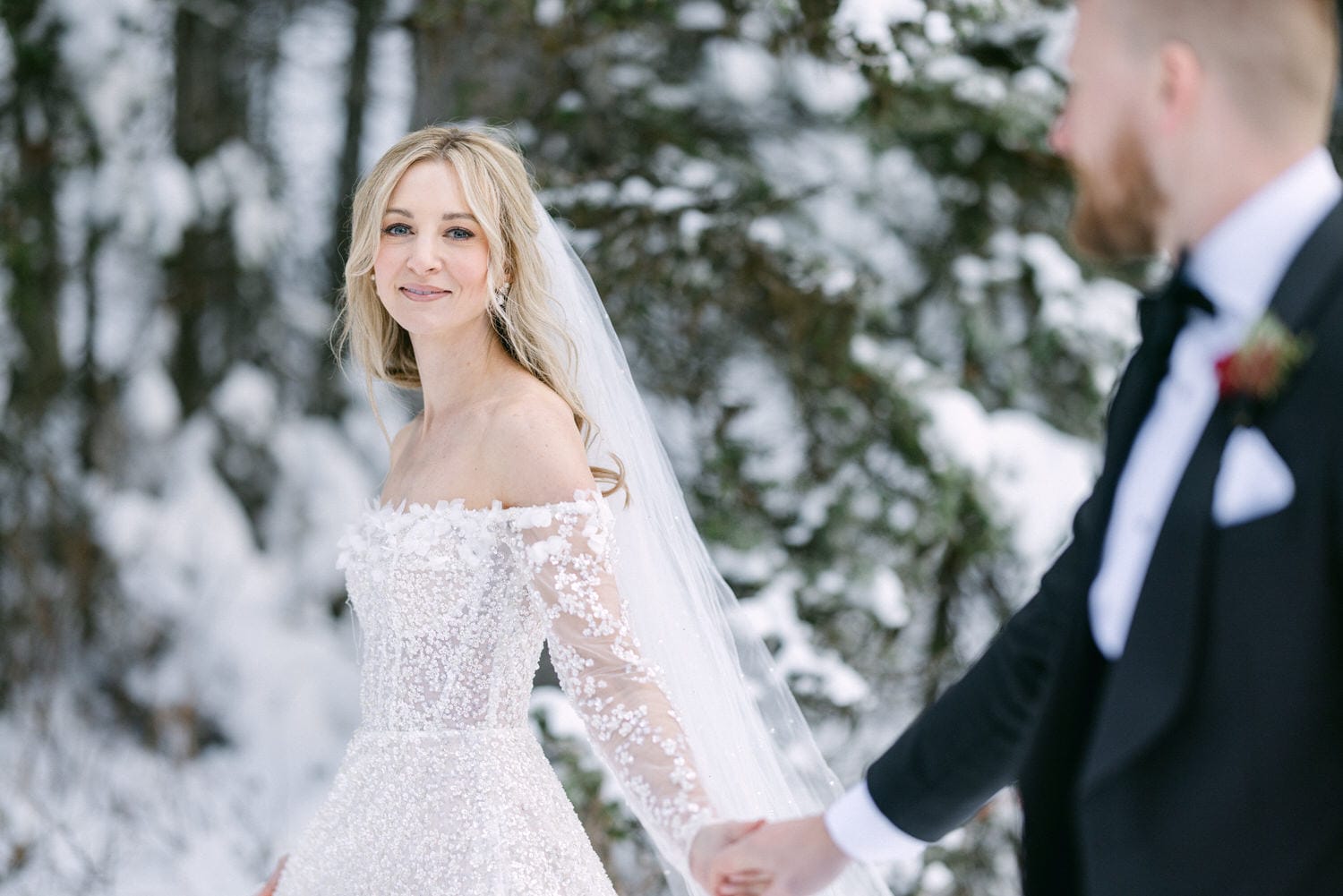 Bride in a white gown smiling at the camera in a snowy forest setting, holding hands with the groom who is partially visible on the right side of the frame.