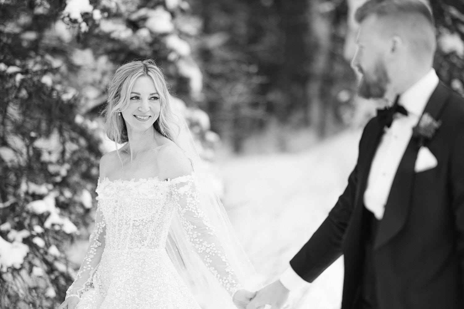 Black and white image of a bride smiling at the camera while holding hands with the groom in a snowy setting.