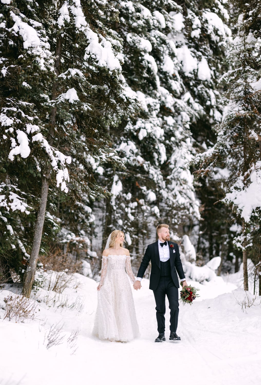 A bride and groom holding hands in a snowy forest setting.