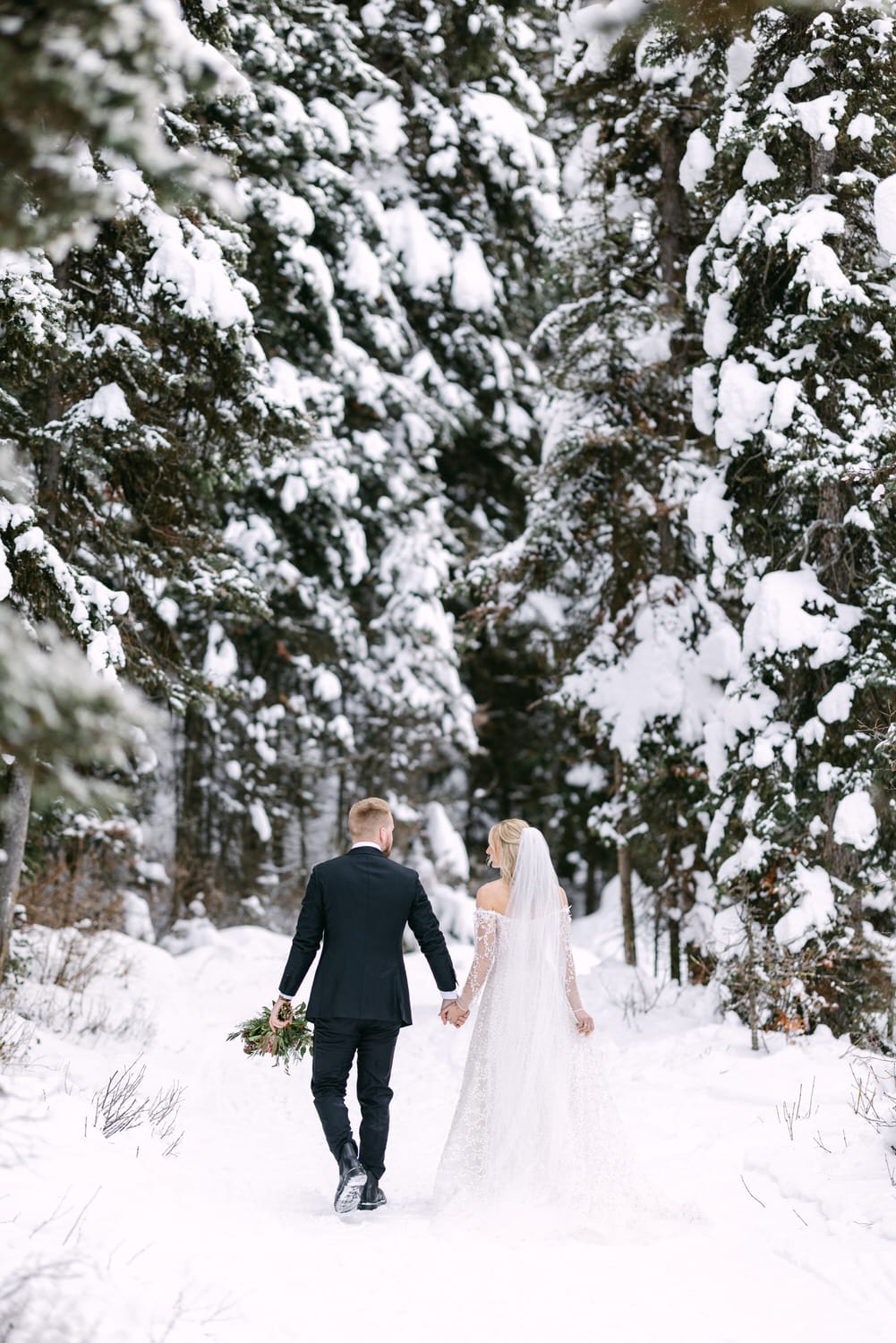 A bride and groom holding hands and walking in a snowy forest pathway, surrounded by trees covered with snow.