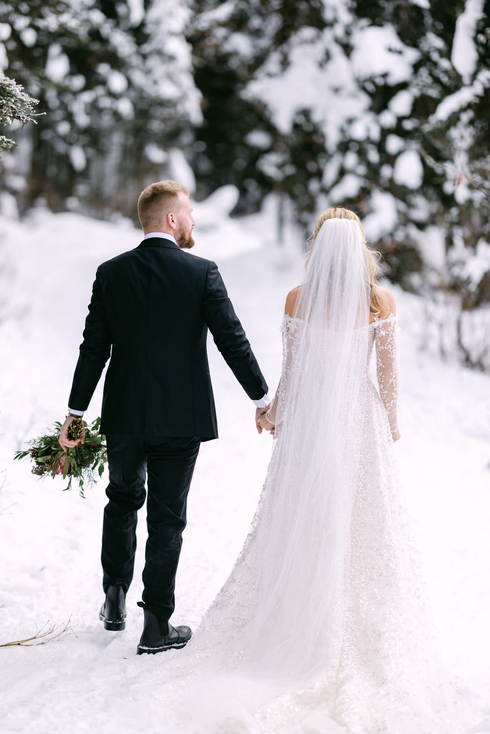 A bride and groom holding hands while walking in a snowy landscape, with the bride in a white wedding dress and veil, and the groom in a black suit.