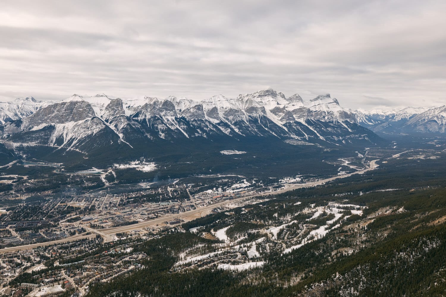 A vast aerial view of a town nestled in a snowy mountain range with a network of roads and forests.