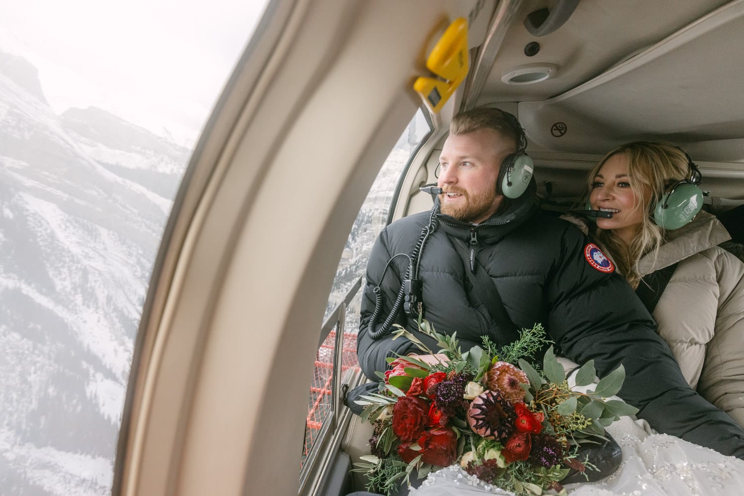 A couple with headsets in a helicopter, the woman holding a bouquet, with snowy mountains visible through the window.