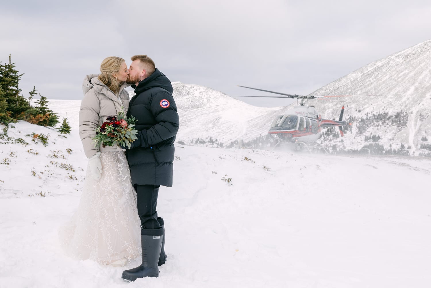 A bride and groom share a kiss in a snowy landscape with a red helicopter landing in the background.