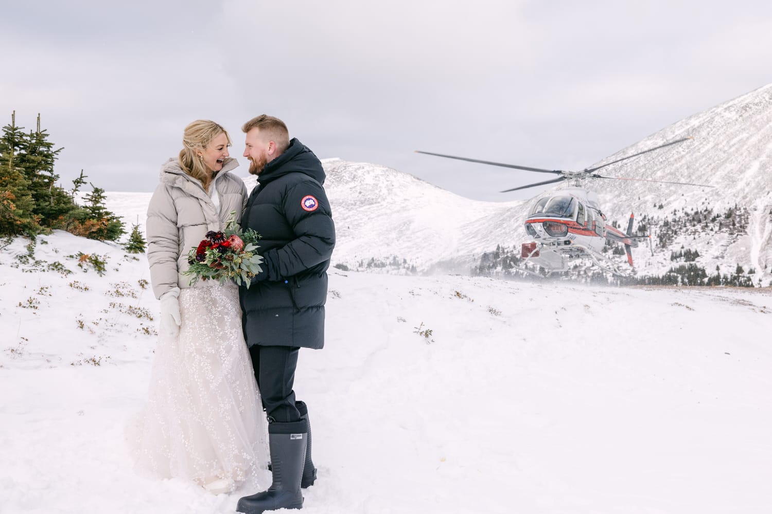 A bride and groom standing together in a snowy landscape with a helicopter in the background.